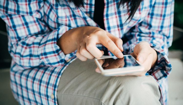A person in a plaid shirt and khakis holds a phone in their left hand while their right index finger taps the screen. 