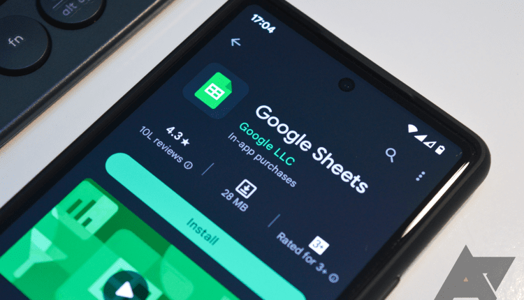 A phone rests on a white desktop. It's display is illuminated and the Google Sheets page in the Play Store is open.