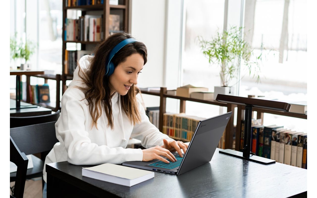 A student wearing headphones sitting at a wooden table with a laptop open in a library setting 