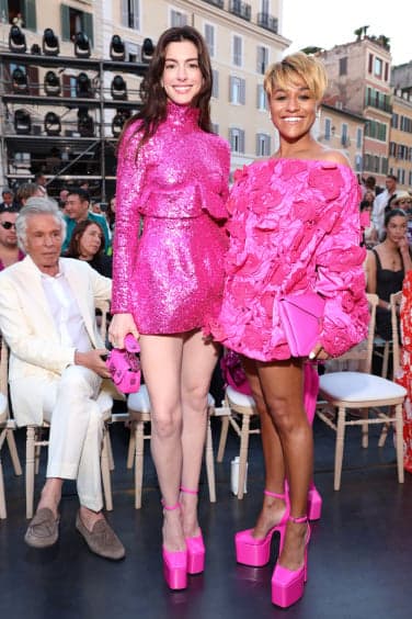 Two women in bright pink outfits stand at a fashion show smiling at the camera.