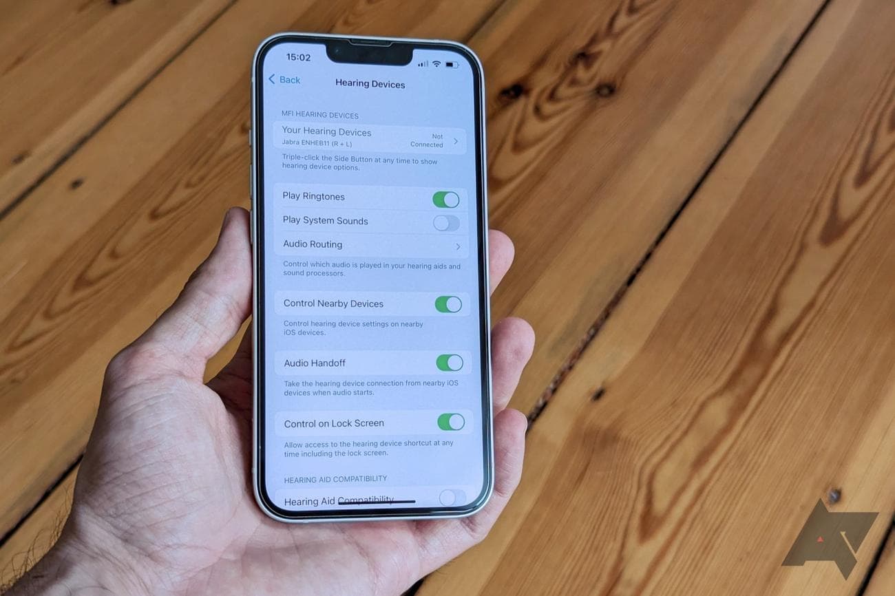 Ένα iPhone κρατιέται από ένα χέρι μπροστά από ένα ξύλινο πάτωμα, εμφανίζοντας την οθόνη ρυθμίσεων των συσκευών ακοής