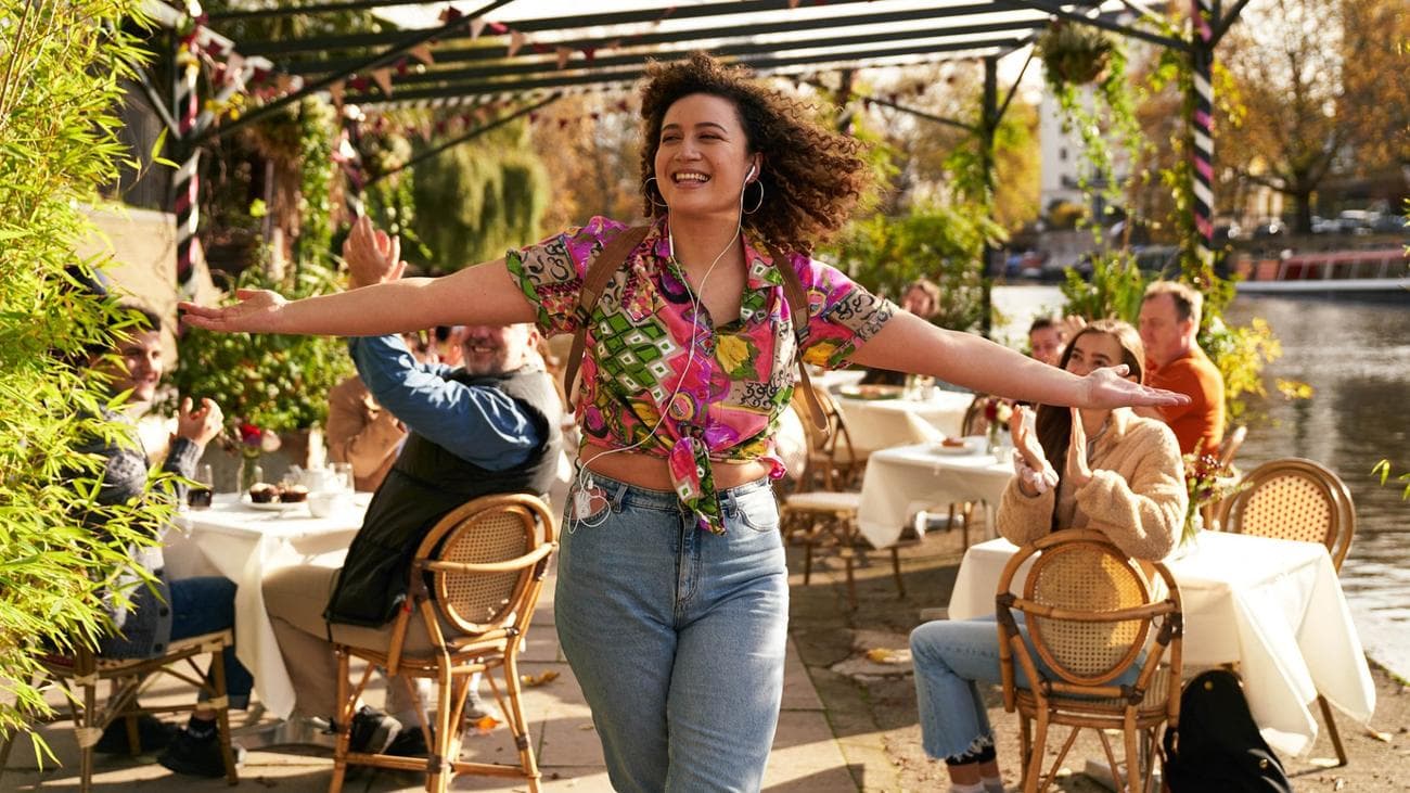 A woman in a colourful blouse smiles and holds her arms out in an outdoor cafe.