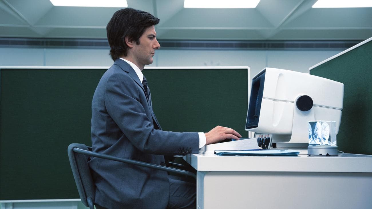 A man in a suit sits in a green cubicle typing at a computer.