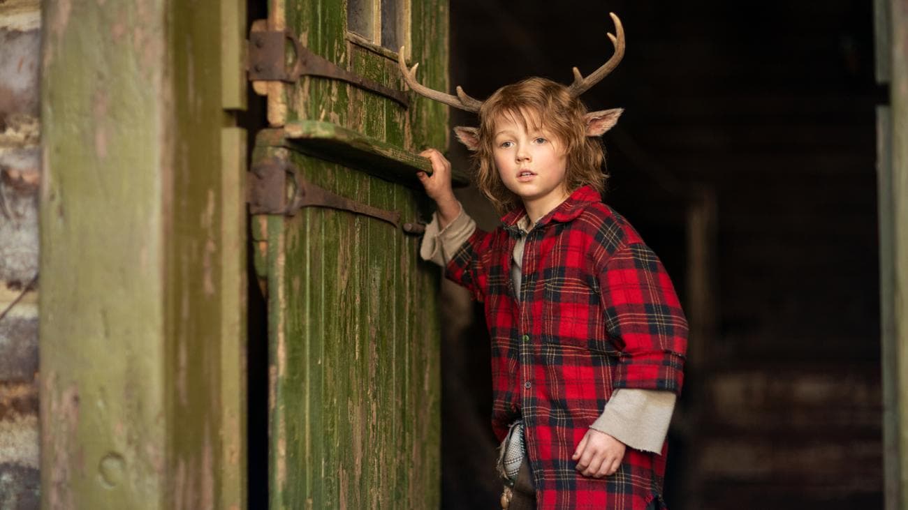 A young boy with antlers wearing a red checked shirt stands at a door.