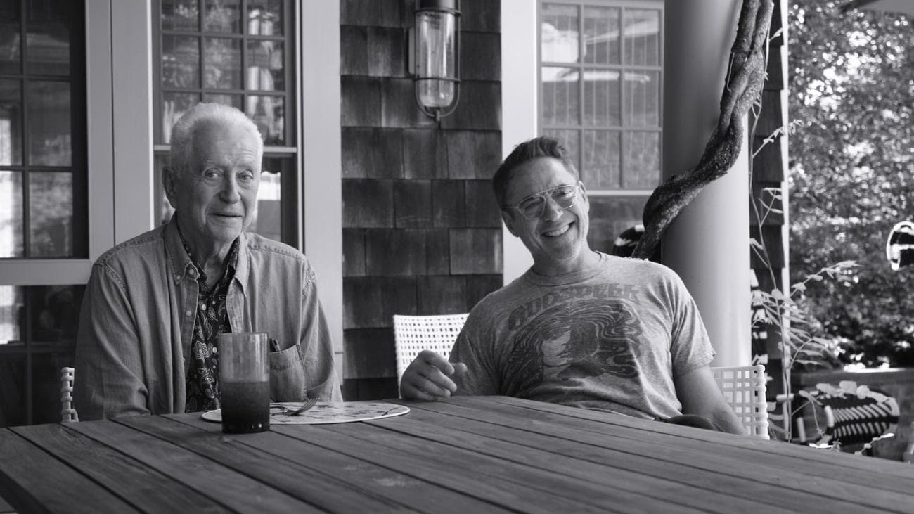 Robert Downey Sr. and Robert Downey Jr. sit together at a table on a porch. 