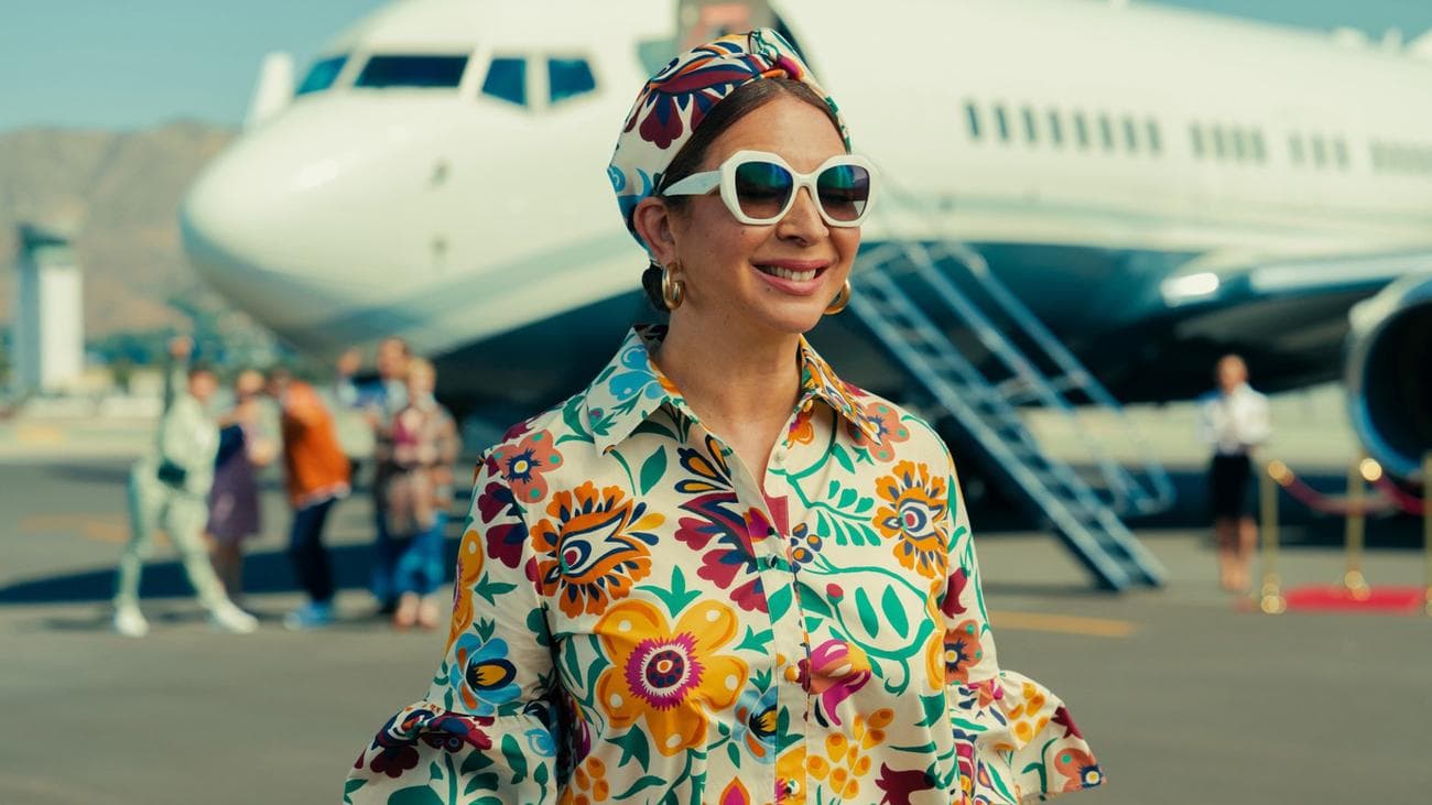 A woman in a bright blouse and sunglasses stands smiling in front of a plane.