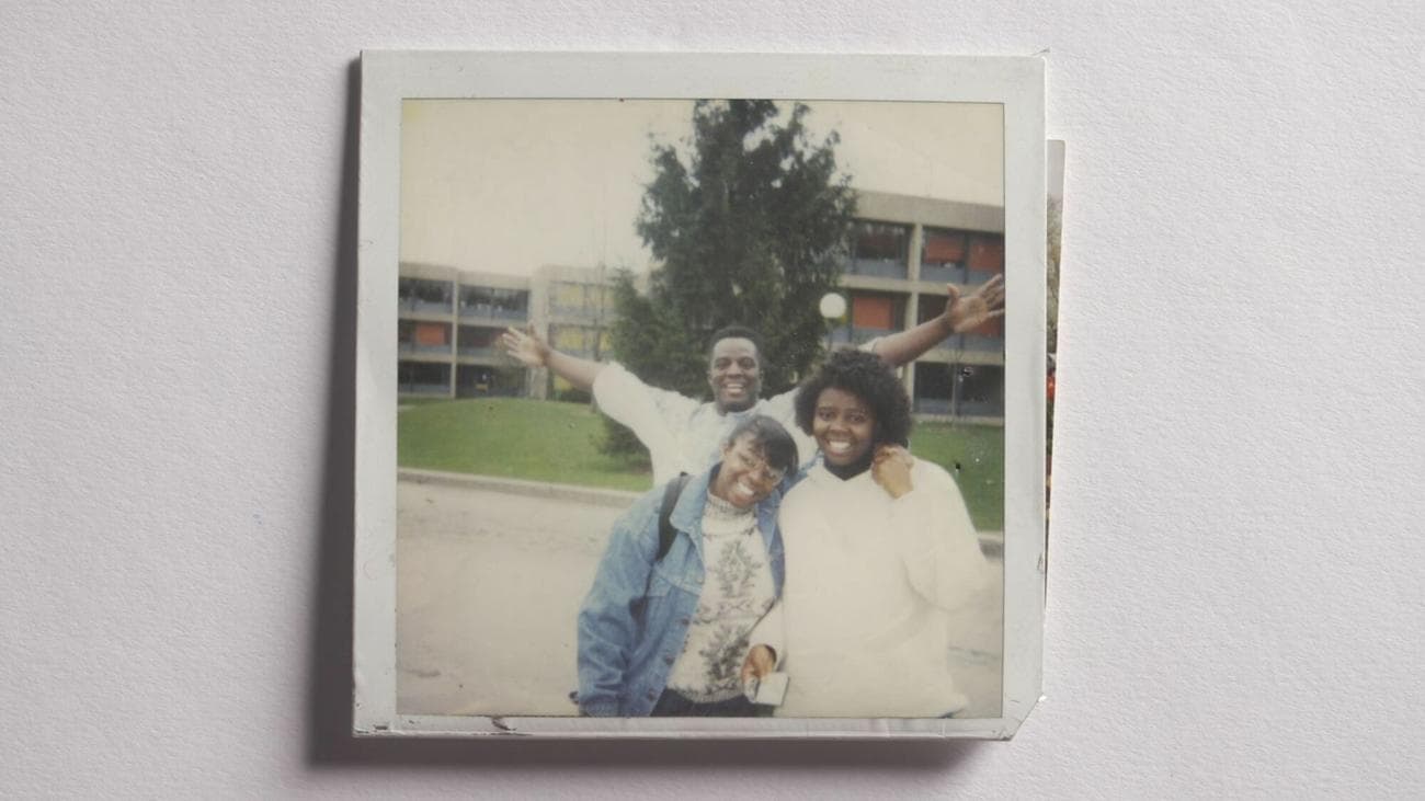 In a faded photograph on a white background, three people stand smiling outside a building.