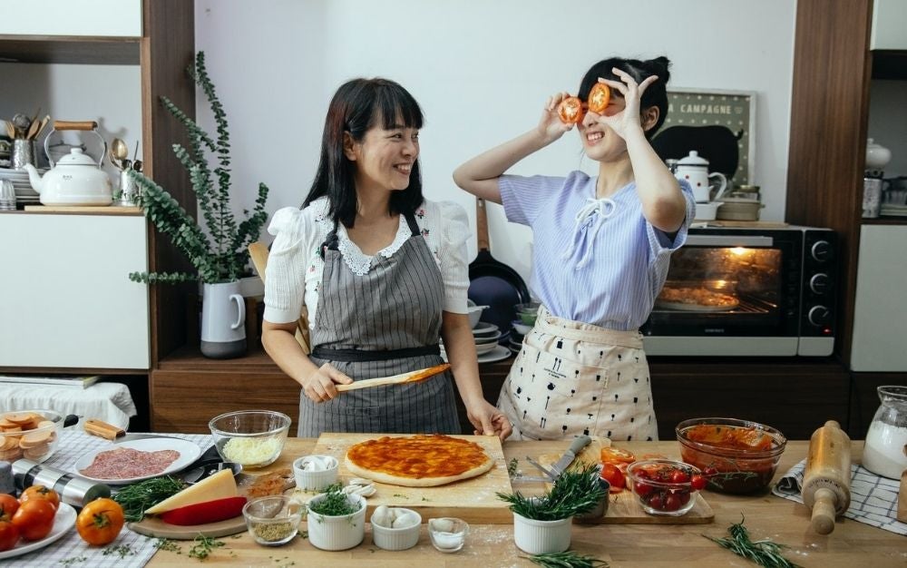 Two woman laughing with each other while cooking pizza in the kitchen.