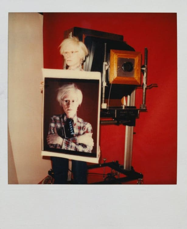 Andy Warhol holds up a large self portrait of himself standing beside a camera.