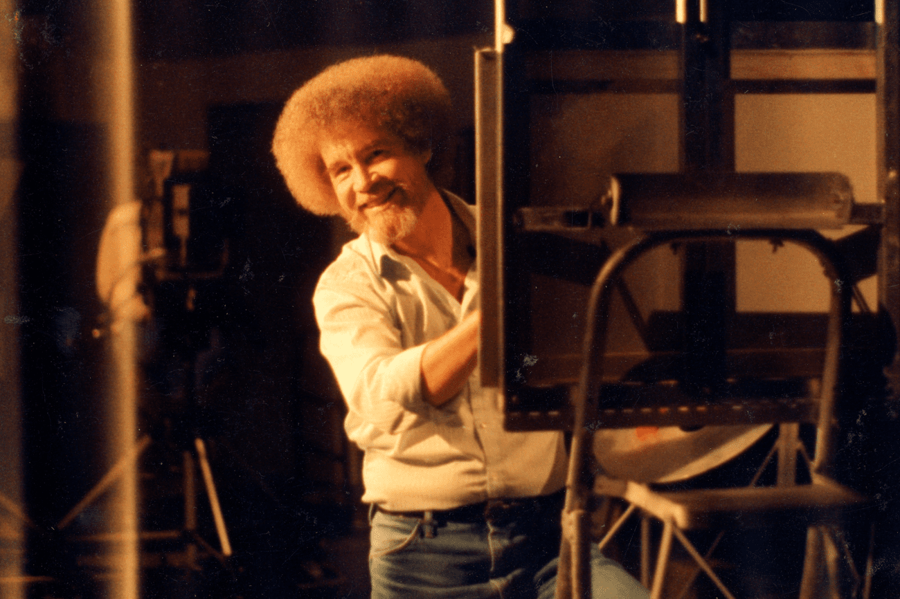 Bob Ross smiling and leaning out from behind an easel.
