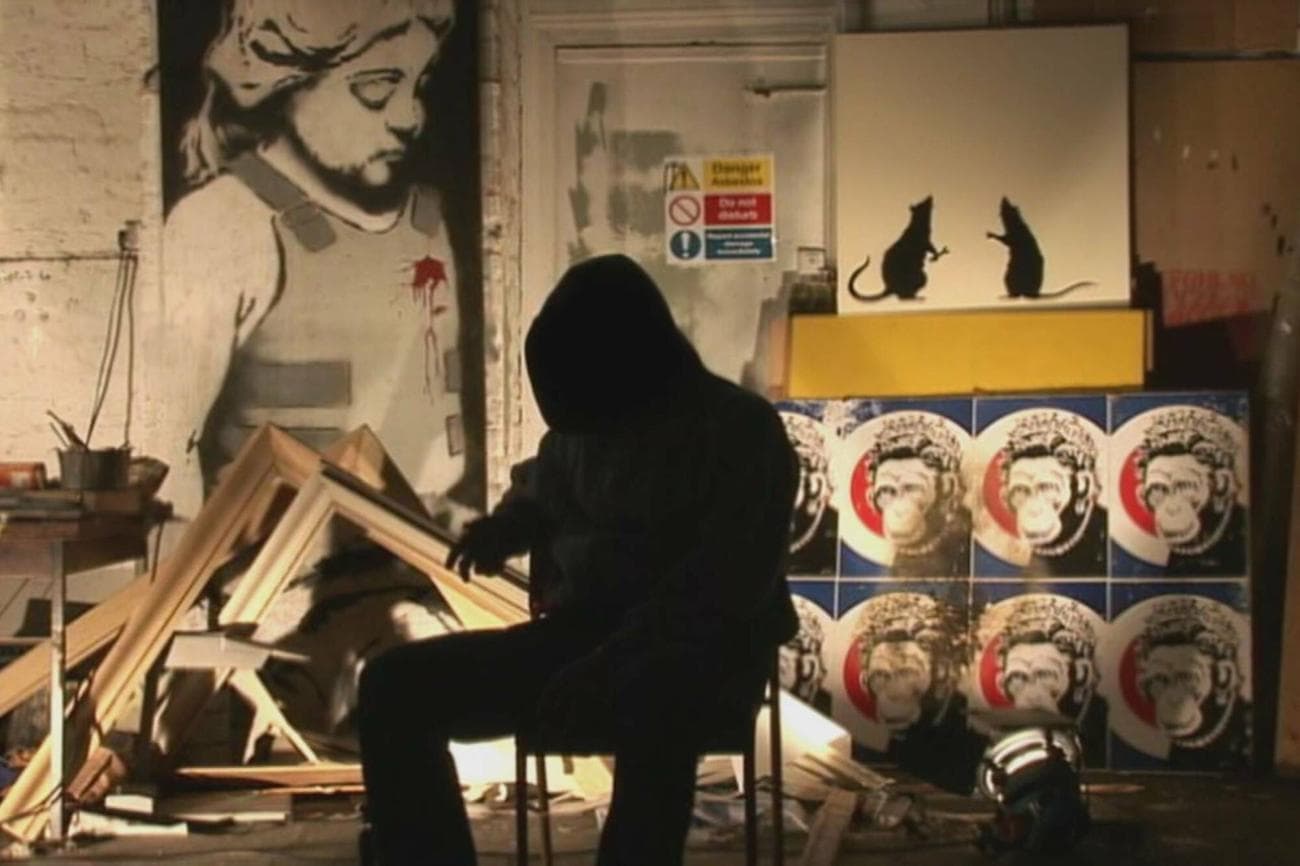 The artist supposedly who is Banksy sits in silhouette in a studio.