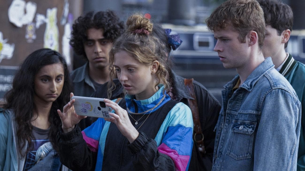 A group of teens gather around a phone looking scared.