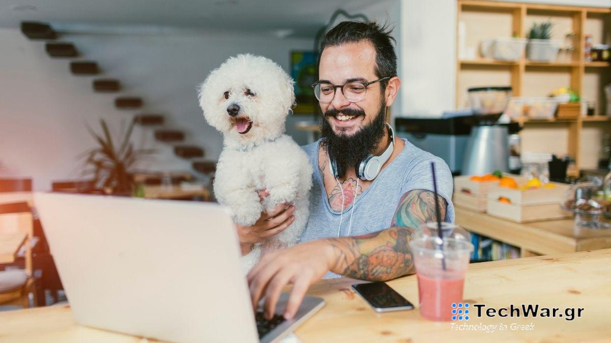 A man holds a small white dog while smiling and looking at a laptop on a table. 