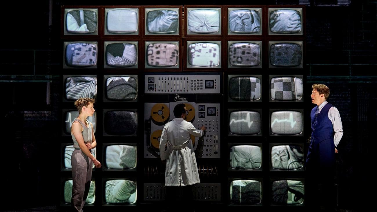 Σε "Stranger Things: The First Shadow" ένας τοίχος από οθόνες χειρίζεται ένας άνδρας με παλτό εργαστηρίου καθώς ένας άντρας και ένα αγόρι κοιτάζονται επίμονα.