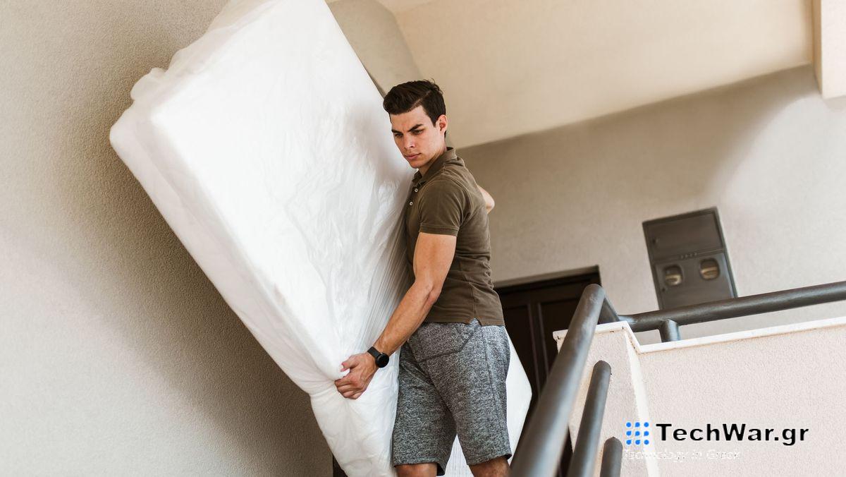 Man in brown shirt and grey shorts carries a white mattress down a flight of stairs