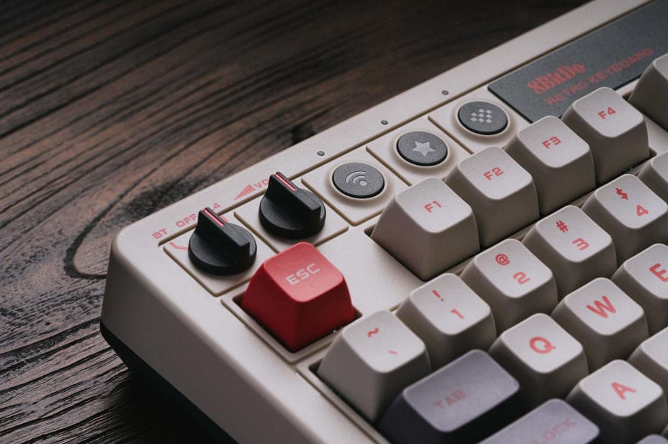 Φωτογραφία προϊόντος του 8BitDo Retro Mechanical Keyboard.  Κοντινό πλάνο του πληκτρολογίου επάνω αριστερά.  Διαθέτει μαύρους διακόπτες και εκρού, κόκκινα ή σκούρα γκρι πλήκτρα.