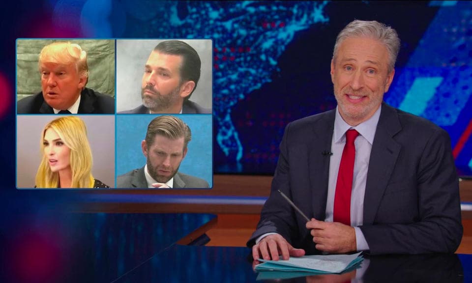 Ακόμα από την επιστροφή του Jon Stewart το 2024 στο The Daily Show.  Ο οικοδεσπότης κάθεται σε ένα γραφείο με μια επικάλυψη που δείχνει φωτογραφίες του Ντόναλντ Τραμπ και των παιδιών του.
