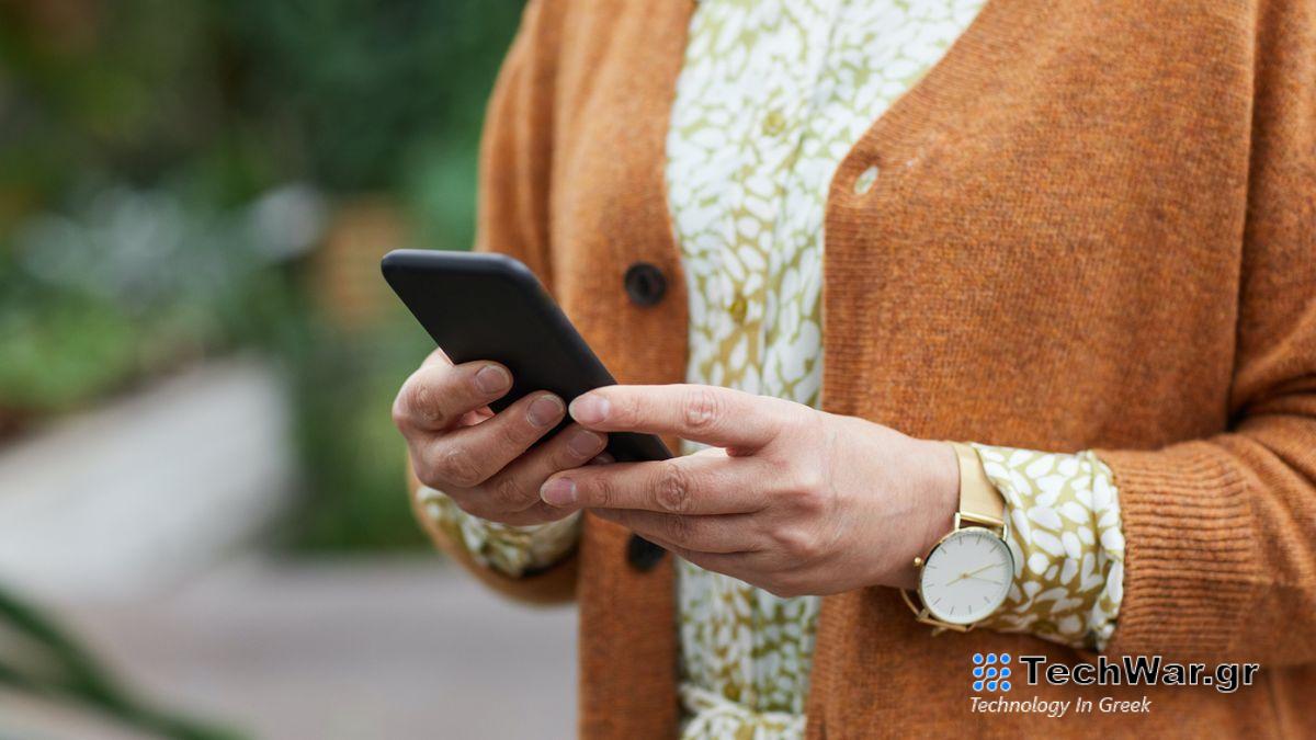 Woman holding phone with brown cardigan and blurred greenery in background